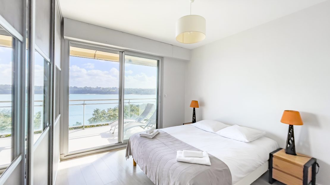 Chambre moderne avec lit, table de nuit et vue sur la mer à travers une grande baie vitrée et un balcon